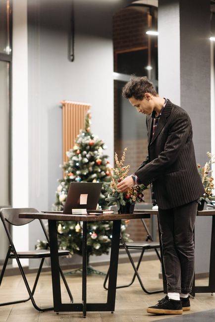 A young man dressed in a dark pinstripe suit is arranging a floral gift atop a wooden table inside a modern, well-lit space. The bouquet features a variety of fresh, seasonal flowers in shades of red,