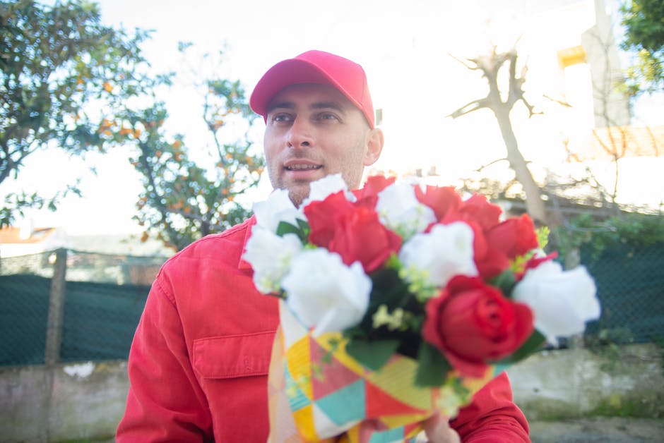 A man in a red cap and matching red jacket is outdoors, holding a vibrant bouquet of fresh flowers wrapped in colorful paper. The bouquet features red roses and white blossoms, with visible green stem