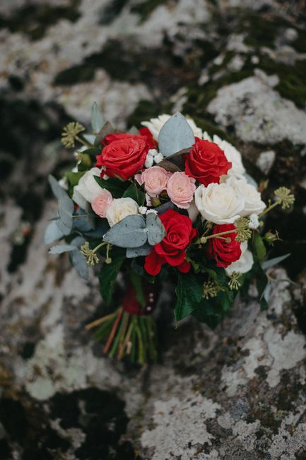 A bouquet of fresh flowers resting on a textured rock surface, arranged with a combination of red roses, white roses, and soft pink ranunculus, complemented by silvery eucalyptus leaves and delicate g