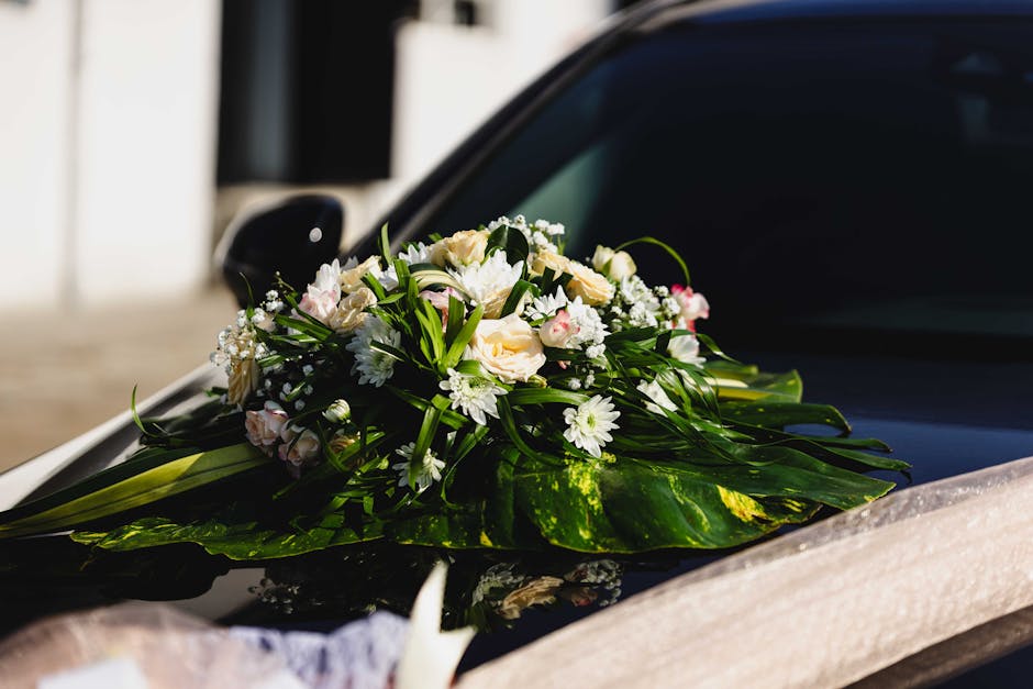 A floral arrangement resting on the hood of a black car, featuring a bouquet of fresh cream and white roses, white daisies, and baby's breath, accented with dark green leaves and wrapped in transparen